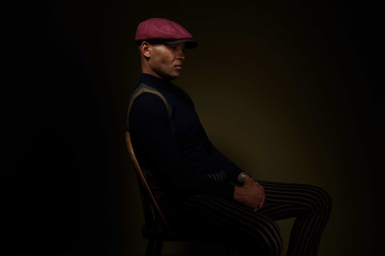 May include: A man wearing a burgundy newsboy cap sits in a chair, wearing a dark blue sweater with a yellow and white stripe pattern and striped pants.