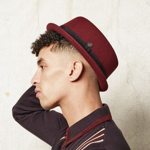 Op de afbeelding: Een bordeauxrode wollen fedora met een zwarte band en een klein knoopdetail. De hoed wordt gedragen door een persoon met kort, krullend haar.