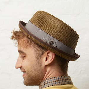 May include: A brown fedora hat with a striped gray band and a dark brown brim. The hat has a small, circular button on the side. The hat is made of woven material.