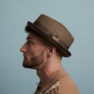 May include: A brown fedora hat with a dark brown band and brim. The hat has a woven texture and a small decorative button. The hat is worn at an angle.