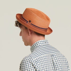 Può includere: Un cappello fedora in paglia intrecciata di colore arancione caldo, con una fascia marrone con una striscia blu navy e un piccolo dettaglio a bottone. Il cappello ha una tesa classica leggermente rialzata. Un accessorio elegante.