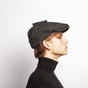 May include: A black and grey herringbone pattern newsboy cap. The cap has a rounded crown and a small, stiff brim. The cap is made of a textured fabric. The person is wearing a black turtleneck.