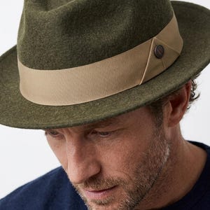 Op de afbeelding: Een olijfgroene vilten fedora-hoed met een brede, gestructureerde rand en een ingedeukte kroon. Een beige grosgrain lint omsluit de basis van de kroon, geaccentueerd met een klein, rond donkerbruin knoopdetail aan de zijkant van de hoedband.