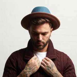 May include: A two-tone fedora hat with a brown brim and a blue crown. The hat is worn by a person wearing a burgundy cable-knit cardigan over a white t-shirt. The person has tattoos on their hands.