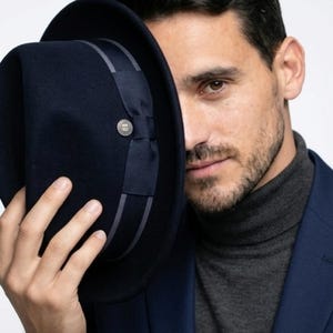 Könnte beinhalten: Ein marineblauer Fedora-Hut mit grauem Band und silbernem Knopfdetail. Der Hut wird hochgehalten und verdeckt teilweise das Gesicht. Die Person trägt einen marineblauen Blazer und einen grauen Rollkragenpullover.