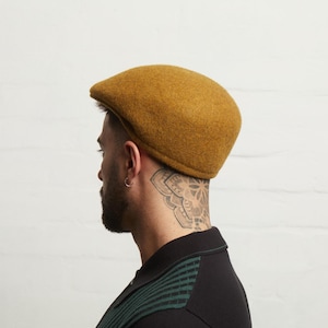 Puede incluir: Gorra plana de color mostaza hecha de tela texturizada. La gorra es de estilo clásico con una corona redondeada y una visera pequeña y rígida. La persona lleva una camisa negra con rayas verdes.