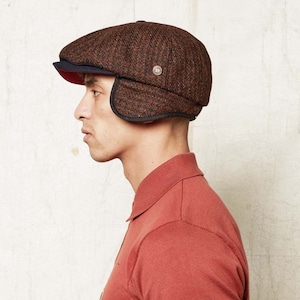 Puede incluir: Una gorra de tweed marrón y negro con orejeras. La gorra tiene una visera negra y un forro rojo.