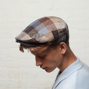 May include: A tweed newsboy cap in a patchwork design, featuring squares of brown, blue, and beige. The cap has a black peak and is made from a textured fabric.