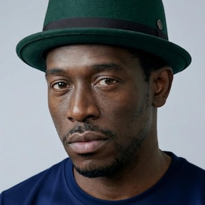 May include: A dark green fedora hat with a black band and a small metal accent. The hat is worn by a person wearing a navy blue top. The hat's brim is slightly curved.