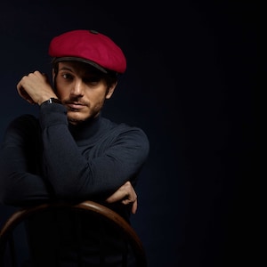 May include: A man wearing a dark blue turtleneck sweater and a red newsboy cap. He is sitting in a wooden chair with his arms crossed.