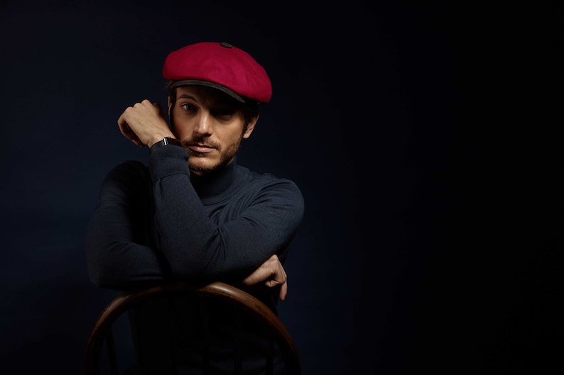 May include: A man wearing a dark blue turtleneck sweater and a red newsboy cap. He is sitting in a wooden chair with his arms crossed.