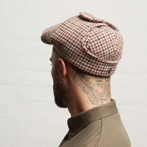 May include: A brown and white houndstooth pattern newsboy cap. The cap has a small brim and a button detail on the side. The cap is made of a textured fabric.