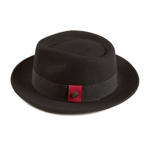 Op de afbeelding: Een zwarte fedora hoed met een zwarte band en een rode accent. De hoed heeft een knoop bovenop.