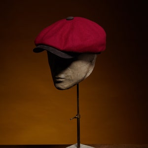 May include: A red wool newsboy cap with a black brim, displayed on a mannequin head on a concrete stand.