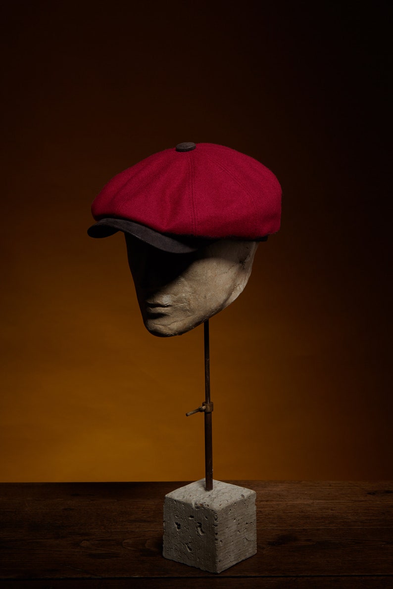 May include: A red wool newsboy cap with a black brim, displayed on a mannequin head on a concrete stand.