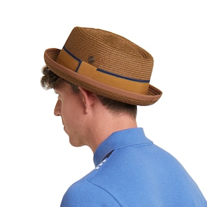 Può includere: Un fedora di paglia marrone con una fascia marrone e blu navy. Il cappello è indossato da una persona con una camicia blu.