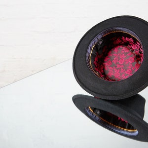 Op de afbeelding: Een zwarte fedora hoed met een gouden rand en een roze bloemenvoering. De hoed is binnenstebuiten gekeerd, waardoor de voering zichtbaar is.