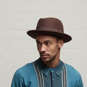 May include: A brown felt fedora hat with a matching brown ribbon band around the crown. The hat features a structured brim and a small metallic emblem detail on the side of the ribbon. The material appears smooth and uniform in texture.