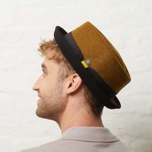 Op de afbeelding: Mosterdkleurige fedora hoed met een zwarte rand en band. De hoed heeft een kleine, ronde knoop en een gele accent. De hoed is schuin geplaatst, wat het ontwerp benadrukt. Een stijlvol accessoire voor elke garderobe.
