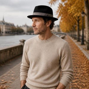 Op de afbeelding: Een zwarte fedora hoed met een grijze lintband gedragen met een beige trui met lange mouwen en ronde hals en een donkergrijze broek. De hoed heeft een gestructureerde kroon en een middelgrote rand, terwijl de trui geribbelde boorden heeft.