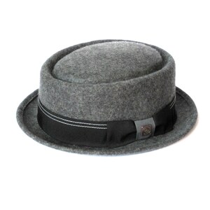 Op de afbeelding: Een grijze wollen fedora hoed met een zwart lint en een klein metalen logo aan de voorkant.