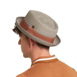 Può includere: Un cappello fedora grigio e marrone con un'ampia tesa. Il cappello è fatto di paglia intrecciata e ha una fascia in pelle marrone attorno alla corona.