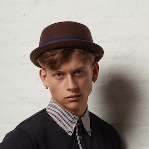 以下が含まれることがあります： 濃い青色のバンドが付いた茶色のボーラーハット。帽子は丸みを帯びたクラウンと短いブリムが付いています。フェルトのような素材で作られています。フォーマルな場面でよく見られるクラシックな帽子スタイルです。周囲：約57cm。