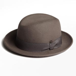 May include: A brown fedora hat with a dark brown ribbon band and a small circular emblem. The hat has a classic, rounded crown and a wide brim. The hat is made of a felt-like material.