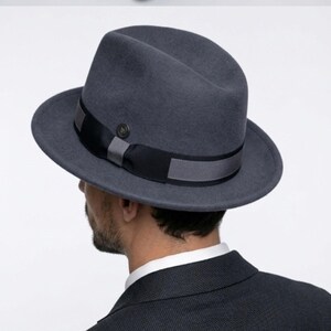May include: A dark grey fedora hat featuring a black and grey band. The hat has a traditional design with a gently creased crown and a broad brim. The hat is made of a felt-like material.