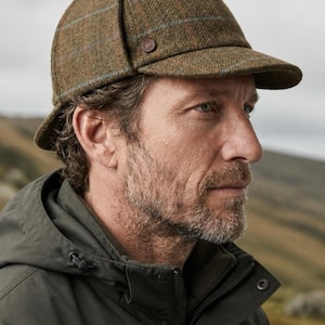 May include: A brown tweed deerstalker hat featuring a subtle blue and orange windowpane check pattern, a short front brim, and a small brown button detail on the side. The hat includes a top tie and ear flaps that are folded up against the crown.