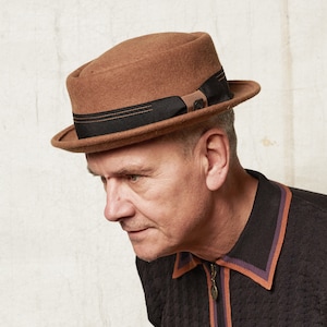 Op de afbeelding: Een bruine wollen fedora-hoed met een zwarte band en een bruin lint. De hoed heeft een platte bovenkant en een rand die naar beneden buigt.