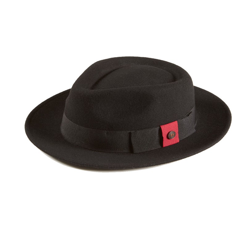 Op de afbeelding: Een zwarte fedora hoed met een rood-zwarte band en een kleine bruine knoop. De hoed heeft een brede rand en een licht ingedeukte kroon.