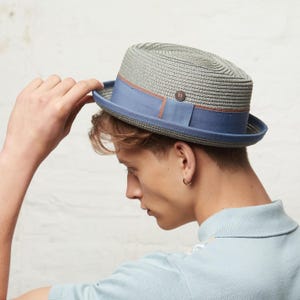 Op de afbeelding: Een grijze fedora hoed met een blauwe band en een bruine accent. De hoed heeft een geweven textuur en een klein knoopdetail. De rand is een donkerdere tint blauw. Een stijlvol accessoire.