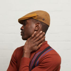 May include: A brown tweed flat cap with a mustard-coloured brim and a small button detail. The cap is worn by a person wearing a rust-coloured polo shirt with blue and black stripes.
