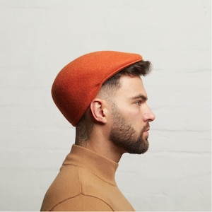 May include: An orange flat cap made from a felt-like material. The cap is a solid colour and has a rounded shape. The cap is worn by a person with a beard and a tattoo on their neck. The person is wearing a tan turtleneck.