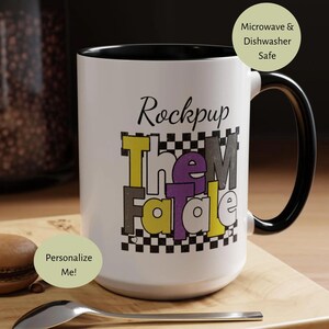 May include: White ceramic mug with a black handle and interior. The mug features the text "Rockpup Them Fatale" in a colorful, retro font. A silver spoon and a macaron are on a wooden surface.