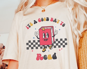 May include: A white t-shirt with a retro-style graphic of a book with a smiling face and the text "It's a good day to read".