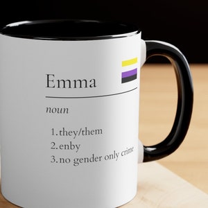 May include: White ceramic mug with a black rim and handle. The mug has the word "Emma" printed on it, followed by the word "noun" and a list of definitions: "1. they/them", "2. enby", "3. no gender only crime". The mug also features a yellow, purple, and black flag.