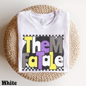 May include: White t-shirt with the text "Them Fatale" in yellow, gray, and purple letters. The text is set against a black and white checkered background. The word "White" is printed at the bottom of the shirt.