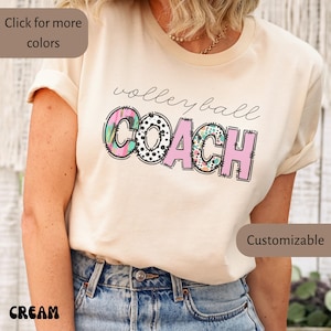 K&ouml;nnte beinhalten: Cremefarbenes T-Shirt mit dem Schriftzug "Volleyball Coach" in einem verspielten Design. Das Wort "Coach" ist in rosa Blockbuchstaben mit bunten Mustern gehalten. Das Shirt ist ein l&auml;ssiger Stil.