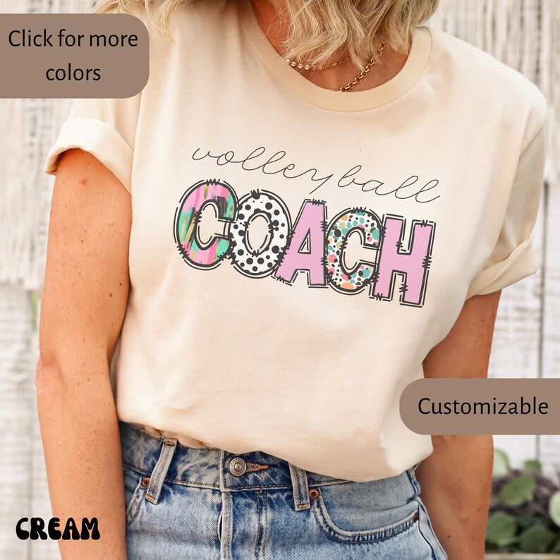 K&ouml;nnte beinhalten: Cremefarbenes T-Shirt mit dem Schriftzug "Volleyball Coach" in einem verspielten Design. Das Wort "Coach" ist in rosa Blockbuchstaben mit bunten Mustern gehalten. Das Shirt ist ein l&auml;ssiger Stil.