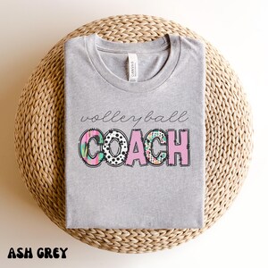 K&ouml;nnte beinhalten: Hellgraues T-Shirt mit dem Schriftzug "volleyball coach" in Schreibschrift. Das Wort "COACH" ist in Blockbuchstaben mit einem farbenfrohen Muster gehalten. Das Shirt wird auf einer gewebten, kreisf&ouml;rmigen Oberfl&auml;che pr&auml;sentiert.