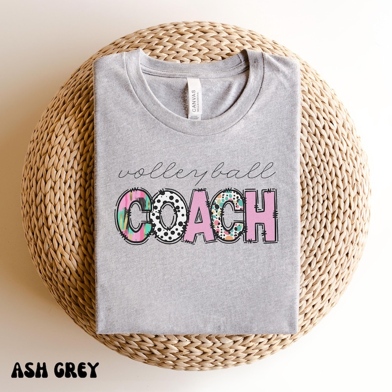 K&ouml;nnte beinhalten: Hellgraues T-Shirt mit dem Schriftzug "volleyball coach" in Schreibschrift. Das Wort "COACH" ist in Blockbuchstaben mit einem farbenfrohen Muster gehalten. Das Shirt wird auf einer gewebten, kreisf&ouml;rmigen Oberfl&auml;che pr&auml;sentiert.