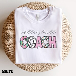 K&ouml;nnte beinhalten: Wei&szlig;es T-Shirt mit dem Aufdruck "volleyball coach". Das Wort "coach" ist in rosa Blockbuchstaben mit schwarzer Umrandung gehalten. Der Buchstabe "O" in "coach" ist mit schwarzen Punkten verziert. Das Wort "volleyball" ist kursiv geschrieben.