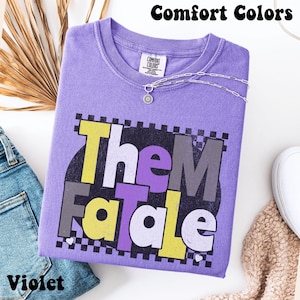 May include: A violet Comfort Colors t-shirt with the text "Them Fatale" in yellow, purple, and gray. The shirt has a crew neck and is accessorized with a silver necklace. The word "Violet" is printed at the bottom left.