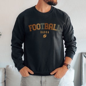 May include: Black crew neck sweatshirt with the text "FOOTBALL DADDA" in orange, green, and brown lettering, with a football graphic below. The sweatshirt is a long-sleeved top.