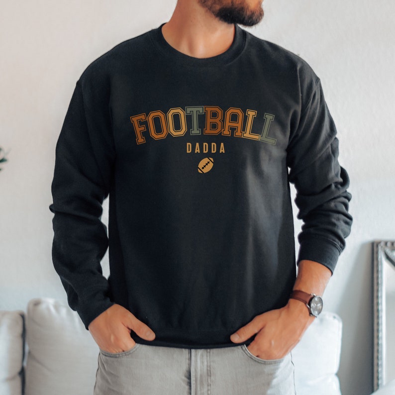 May include: Black crew neck sweatshirt with the text "FOOTBALL DADDA" in orange, green, and brown lettering, with a football graphic below. The sweatshirt is a long-sleeved top.