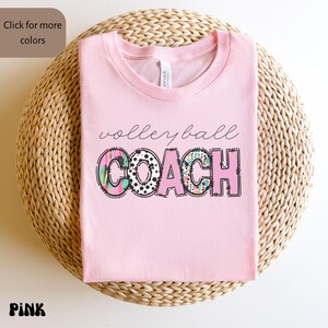 K&ouml;nnte beinhalten: Ein rosa T-Shirt mit dem Aufdruck "volleyball COACH". Das Wort "volleyball" ist kursiv geschrieben, w&auml;hrend "COACH" in fetten Blockbuchstaben mit bunten Mustern gehalten ist. Das T-Shirt liegt gefaltet auf einer gewebten Oberfl&auml;che.