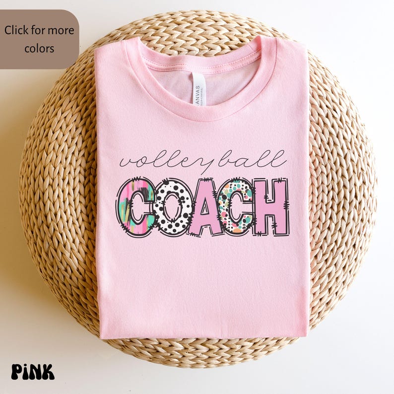 K&ouml;nnte beinhalten: Ein rosa T-Shirt mit dem Aufdruck "volleyball COACH". Das Wort "volleyball" ist kursiv geschrieben, w&auml;hrend "COACH" in fetten Blockbuchstaben mit bunten Mustern gehalten ist. Das T-Shirt liegt gefaltet auf einer gewebten Oberfl&auml;che.
