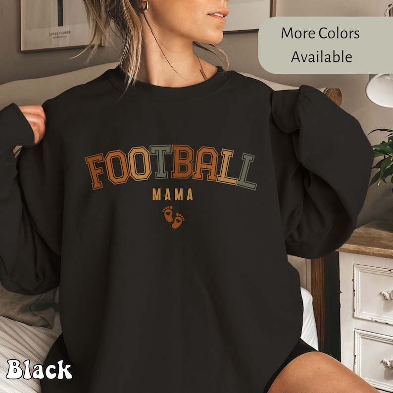 May include: Black sweatshirt with the text "FOOTBALL MAMA" in orange, yellow, and green lettering, with two small orange footprints below. The words "More Colors Available" are in a light gray box.